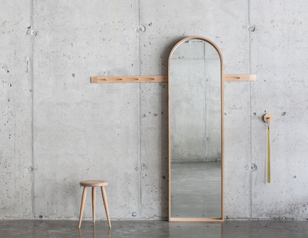a concrete wall with a light oak wood rail with brass pegs, a full length arched mirror, a three-legged stool and a brass shoe horn.