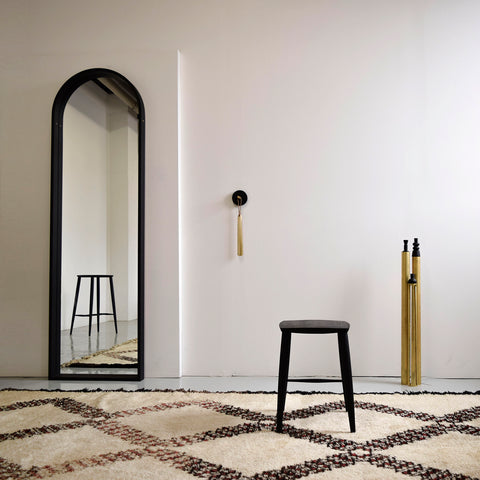 coolican and company showroom, featuring an arched full-length mirror, black minimal stools, brass shoehorn and brass decor.  A Moroccan rug on the floor.